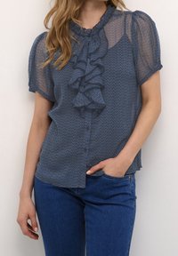 Blå, mönstrad genomskinlig blus med kort puffärmar, volang framtill, rundad hals och knappstängning, kombinerad med denimjeans.