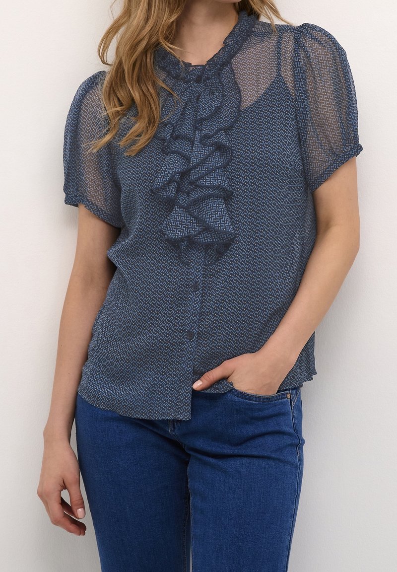 Blå, mönstrad genomskinlig blus med kort puffärmar, volang framtill, rundad hals och knappstängning, kombinerad med denimjeans.