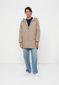Beige hoodie met een ritssluiting aan de voorkant en een kleurrijke streep; gecombineerd met lichtblauwe wijde jeans en witte sneakers. Glad textuur.