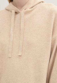 Felpa beige realizzata in materiale morbido, con una vestibilità comoda, cappuccio con coulisse e due linee verticali dove si attaccano le coulisse.