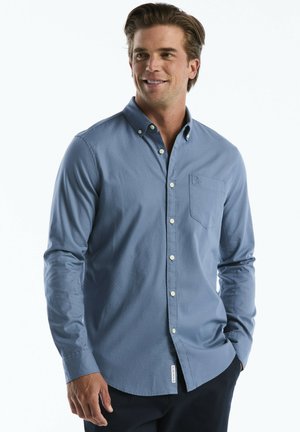 Man wearing a light blue button-up long-sleeve shirt standing with one hand in pocket, smiling and looking to the side.