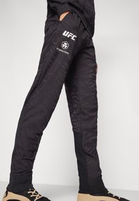 Sorte athletic bukser med et tekstureret tigerstribe mønster, prydet med UFC-logoet og crypto.com branding i hvid. Elastiske manchetter ved anklerne.