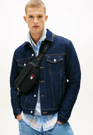 Jeune homme aux cheveux courts portant une veste en denim foncé, une chemise à carreaux bleu clair et un sac bandoulière noir avec le logo "Tommy Jeans".
