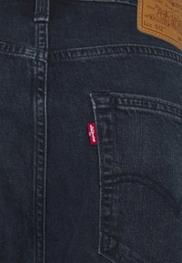 Tmavě modré džíny z denimu s zadní kapsou, na které je červená nášivka Levi's a béžová kožená nášivka, ukazující klasické prošívání a strukturu.