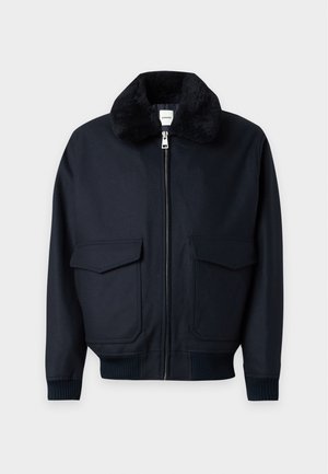 Männliches Model trägt schwarze Bomberjacke mit Fellkragen, lockere schwarze Hose, weißes Hemd und schwarz-weiße Schuhe, steht vor weißem Hintergrund.
