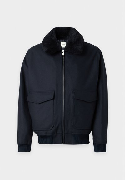 Veste noire zippée avec col doux et pelucheux, deux grandes poches avant à rabat, poignets et ceinture côtelés.