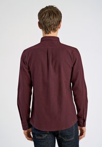 Bordeaux knoopshirt van een gestructureerde stof, lange mouwen, een kraag en een afgeronde zoom. Voorzien van een rugplooi en zwarte knopen.