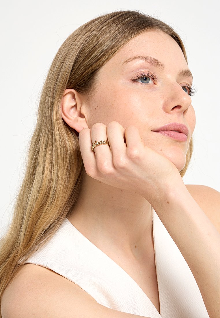 Frau mit langen blonden Haaren, die ihr Kinn auf ihre Hand stützt, trägt ein weißes ärmelloses Top und einen goldenen Kettenring an ihrem Finger.