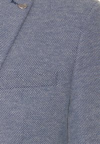 Blå och vit texturerad jacktyg med en närbild av en liten bröstficka och en silverfärgad bladformad pin på slaget.