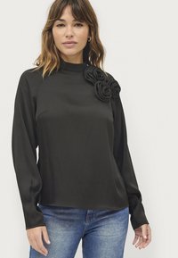 Zwarte lange mouw top met een hoge hals en textuur bloemenaccenten op één schouder, gemaakt van een gladde stof, gecombineerd met een blauwe spijkerbroek.