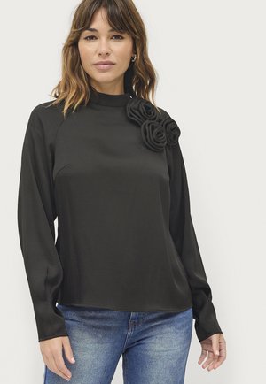 Schwarzes Langarmoberteil mit hohem Kragen und strukturierten floralen Akzenten auf einer Schulter, gefertigt aus einem glatten Stoff, kombiniert mit blauen Jeans.
