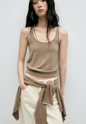 Junge Frau mit langen schwarzen Haaren trägt ein braunes ärmelloses Oberteil und beige Hosen mit einem braunen Pullover um die Taille gebunden.