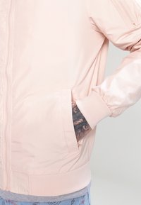 Světle růžová bomber bunda vyrobená z hladké tkaniny, s zipovým zapínáním, boční kapsou a ribbovanými manžetami. Na zápěstí jsou viditelné dekorativní tetování.