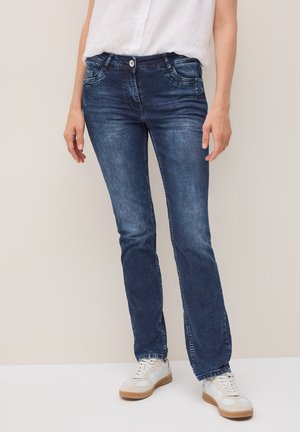 Une personne se tient debout, portant un jean bleu foncé et des baskets blanches, la main sur la hanche, devant un fond neutre.