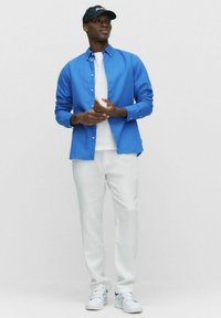 Blauwe overhemd met knopen over een wit T-shirt, gecombineerd met witte broeken en witte sneakers met groene accenten. Model staat tegen een neutrale achtergrond.