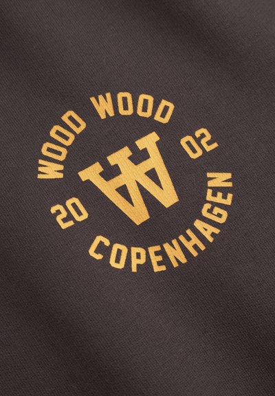 Texte jaune "WOOD WOOD 2002 COPENHAGUE" et logo stylisé avec les lettres "W" et "A" se chevauchant sur un tissu sombre.