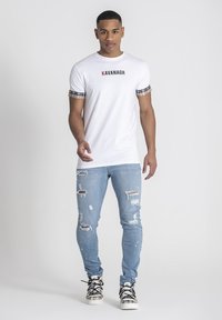 Camiseta blanca con el logo "KAVANAGH" en negro y rojo, mangas de patrón a cuadros, combinada con jeans skinny descoloridos de color azul claro y zapatillas negras.