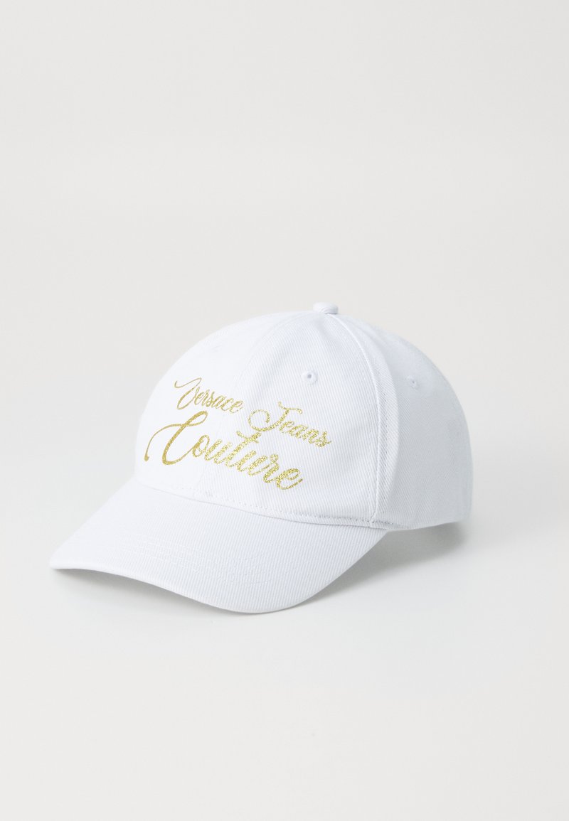 White baseball cap made of cotton with a curved brim. Features gold embroidered text "Versace Jeans Couture" on the front.