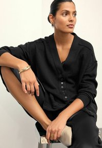 Melna pārmērīga izmēra kardigāns ar V veida kakla izgriezumu un pogu priekšpusi, kombinēts ar melnām biksēm. Gluda tekstūra un brīvs piegriezums ar minimālu apdari.