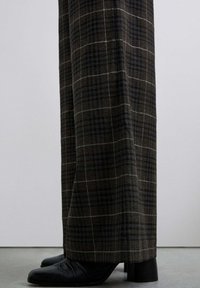 Pantalons larges en tissu à carreaux foncés présentant des nuances de brun et de gris. Texture lisse avec un léger éclat, associés à des chaussures noires.