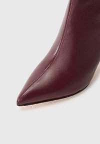 Botte à bout pointu en cuir bordeaux avec une texture lisse et un design élégant, dotée d'un talon haut et de détails de couture minimalistes.