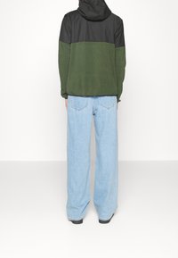 Bluza z kapturem w czarnym górnym i zielonym dolnym odcieniu, zestawiona z jasnoniebieskimi szerokimi spodniami dżinsowymi. Posiada teksturowaną tkaninę i luźny krój.