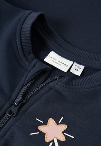 Chaqueta azul marino con cremallera y diseño de estrella con detalles dorados y rayos blancos. Fabricada con un tejido suave y con cuello y puños acanalados.