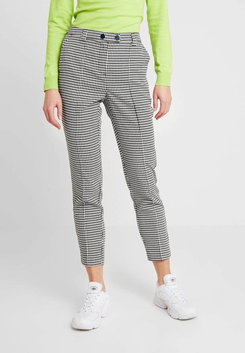 Personne portant un pantalon court motif pied-de-poule noir et blanc, des baskets blanches et un haut manches longues vert néon, debout sur un sol blanc.