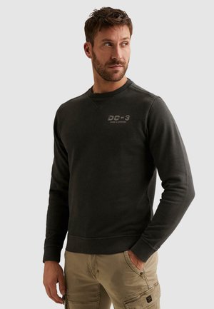 Mann trägt einen dunkelgrauen Langarm-Sweatshirt mit dem Text „DC-3 PME Legend“ und beigefarbenen Cargohosen, schaut nach links mit einer Hand in der Tasche.