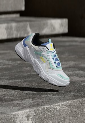 White, blue, and mint green FILA sneaker with yellow logo, suspended above concrete steps casting a shadow beneath.