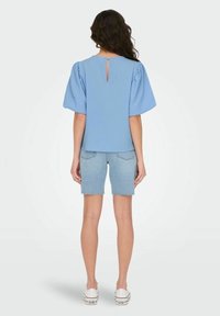 Top corto a maniche corte blu con texture e dettaglio a chiave sulla schiena, abbinato a pantaloncini di jeans chiari e sneakers bianche.