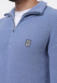 Pull à zip bleu clair avec une texture côtelée, présentant un emblème de blason noir et blanc sur la poitrine gauche.