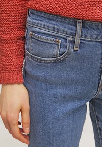 Blaue Jeans aus Denim mit klassischer Passform, ausgestattet mit einer Gesäßtasche und gesteppten Details. Kombiniert mit einem strukturierten Korallfarbenen Strickpullover.