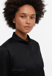Black satin blouse with a tall, standing collar and long sleeves. Smooth texture with a minimalist design, featuring a hidden button placket.