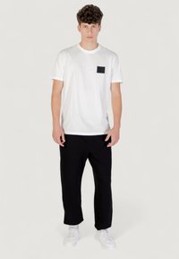 Witte katoenen T-shirt met korte mouwen en een zwarte borstzak, gecombineerd met een zwarte broek en witte sneakers, tegen een neutrale achtergrond.