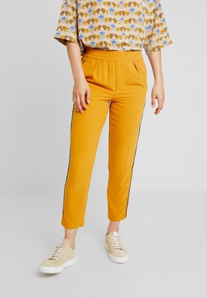 Persoon die mosterdgele taps toelopende broek met zwarte zijstrepen draagt, een beige top met dierenprint en beige sneakers op een grijze vloer.