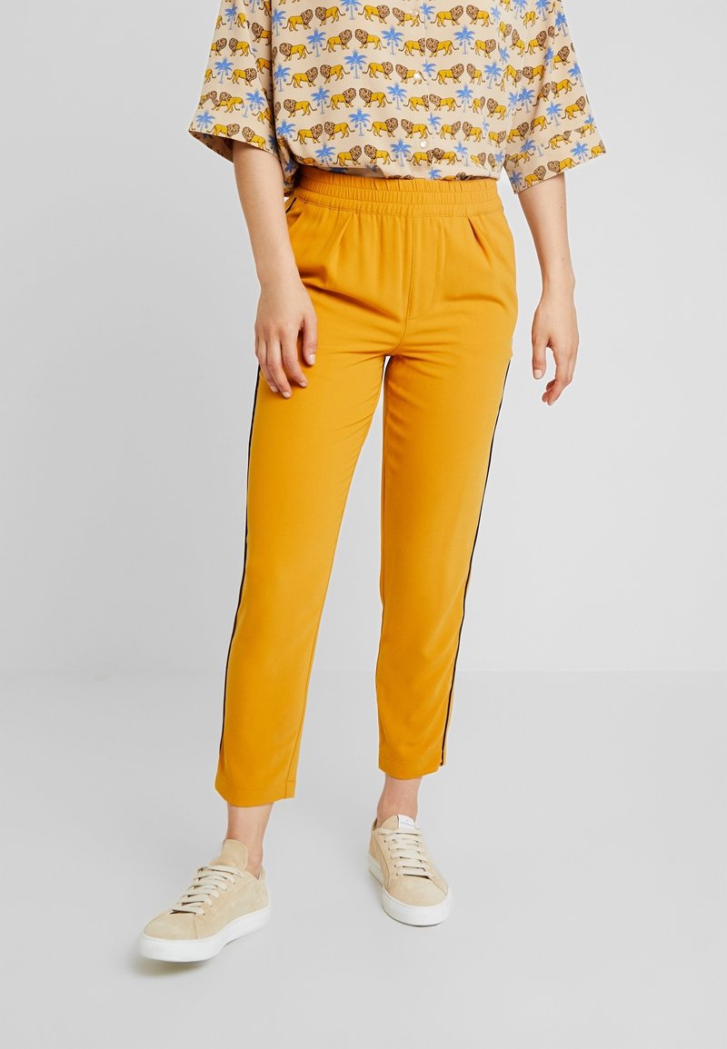 Personne portant un pantalon étroit jaune moutarde avec des bandes noires sur les côtés, un haut beige à imprimé animalier et des baskets beiges sur un sol gris.