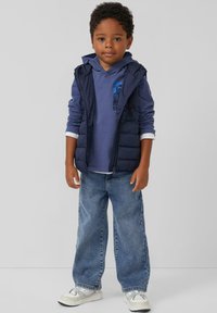 Marine blauwe puffer vest over een blauwe hoodie, lichtblauwe jeans en witte sneakers. De hoodie heeft een grafische print; casual, gelaagde look.