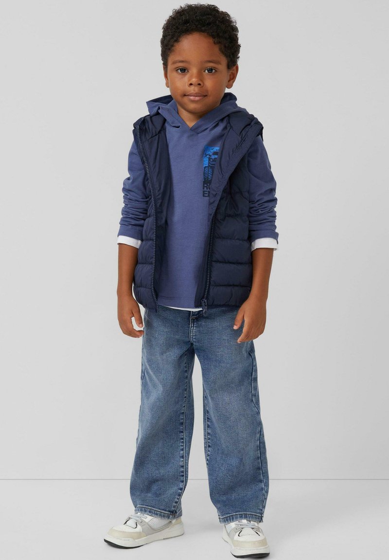 Marine blauwe puffer vest over een blauwe hoodie, lichtblauwe jeans en witte sneakers. De hoodie heeft een grafische print; casual, gelaagde look.