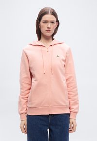 Νεαρός που φοράει ροζ παστέλ φούτερ με φερμουάρ και μικρό πράσινο λογότυπο στο στήθος, καθώς και μπλε τζιν, στέκεται μπροστά σε απλό ανοιχτό φόντο.