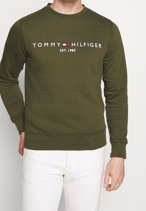 Hombre vistiendo una sudadera de manga larga verde oliva con el texto "Tommy Hilfiger EST. 1985" y el logo, combinada con pantalones blancos.