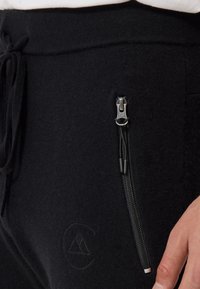 Schwarze Jogginghosen aus weichem Stoff mit einer seitlichen Reißverschlusstasche. Merkmale ein geprägtes Logo und einen verstellbaren Kordelzugbund.