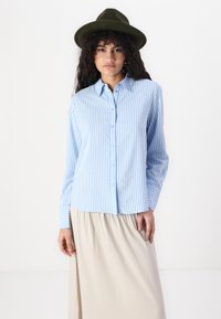 Light blue striped button-up shirt with long sleeves and cuffs, paired with a beige skirt. Model wearing an olive green hat.