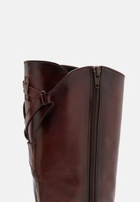 Bota de cuero marrón hasta la rodilla con cierre de cremallera, borde superior curvado y detalle de correa decorativa en el lateral. Textura suave y acabado pulido.