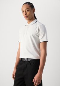Helles beige Polohemd mit dunkelblauem Kragen und Ärmelabschlüssen, ausgestattet mit einer Knopfleiste. Kombiniert mit schwarzen Jeans und einem schwarzen Gürtel.