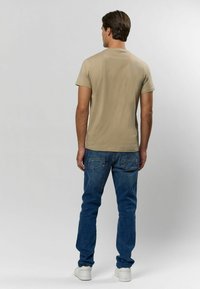 Beige tričko s krátkým rukávem a kulatým výstřihem, zkombinované s modrými džíny a bílými teniskami. Zobrazeno z boku.