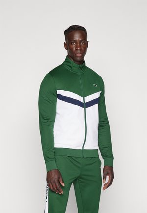 Chaqueta verde y blanca con cremallera y un alto cuello, con un patrón de franjas en color marino. Combinada con pantalones verdes a juego, mostrando el logo de la marca.