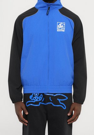 Mand iført en blå og sort lynlåstrøje med et "icecream"-logo og sorte bukser med en blå grafik af en løbende skabning.