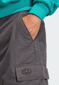 Sivé cargo šortky s viditeľnými bočnými vreckami, textúrovanou látkou a čiernou oválnou záplatou s rastlinným motívom na spodnom vrecku.