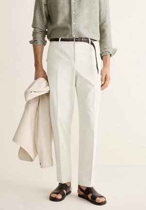 Hombre que lleva camisa de manga larga remangada, pantalones blancos con cinturón trenzado marrón, sostiene una chaqueta beige y lleva sandalias de cuero negras.